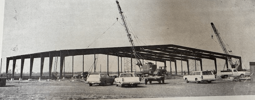 Black and white photo showing the steel framework of BonnaVilla’s first plant under construction in Aurora, Nebraska, with cranes and trucks on-site.