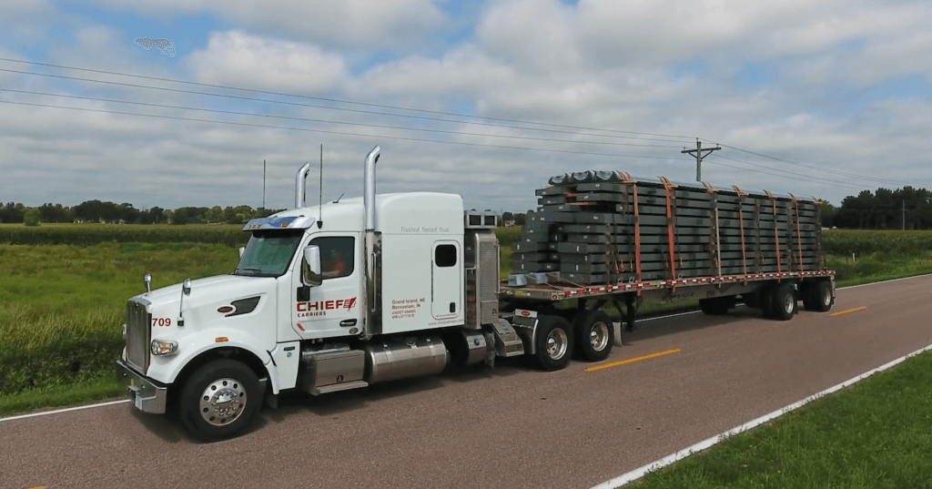A white Chief Carriers semi-truck hauling a flatbed trailer loaded with steel components, driving along a rural highway surrounded by fields under a cloudy sky.