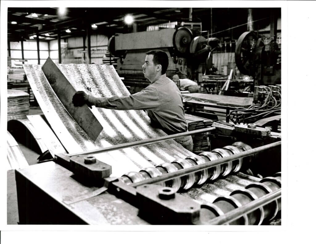 Black and white photo of a worker handling a corrugated steel panel in a manufacturing plant.