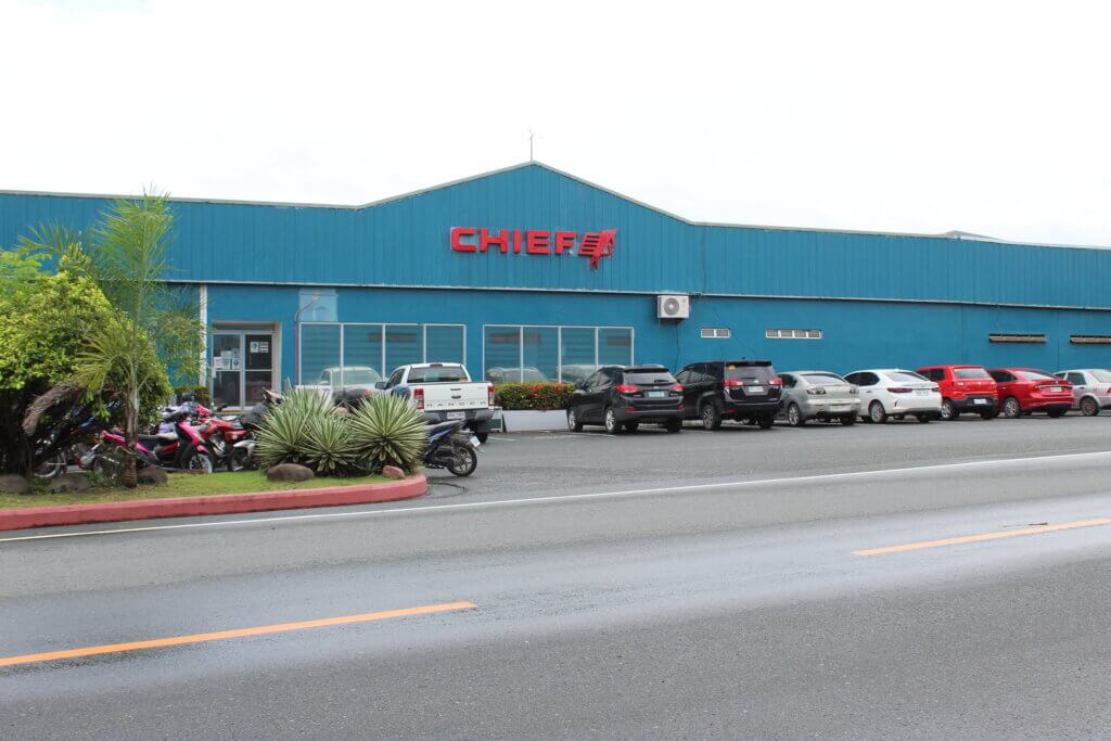Color photo of Chief Agri’s bright blue facility in the Philippines, with the red Chief logo on the front and several cars and motorcycles parked outside.